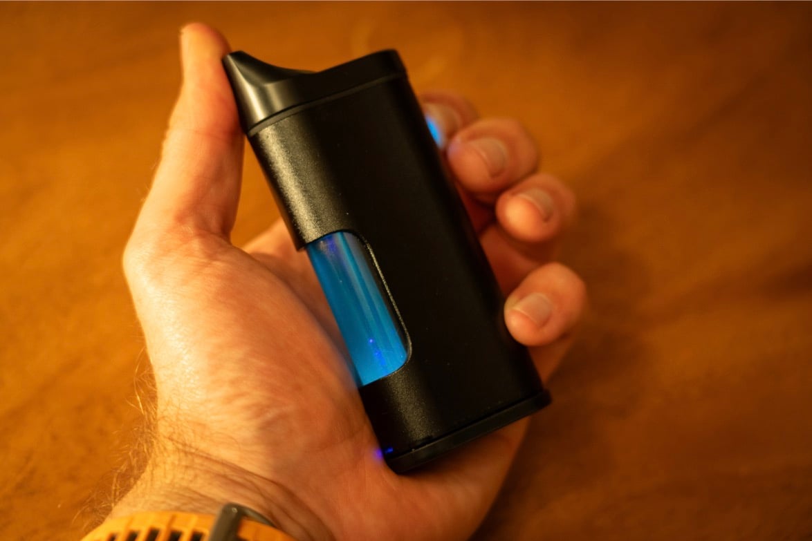 An image of a hand holding a black vape with a vibrant blue chamber where you can faintly see a laser.