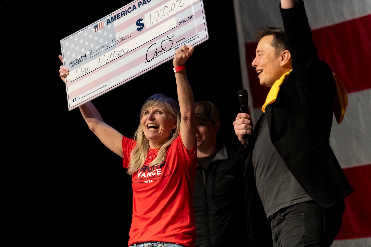 A woman stands next to Elon Musk, holding up a giant check.